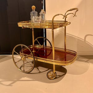 Könnte beinhalten: Ein Barwagen im Vintage-Stil mit goldfarbenem Rahmen und zwei Ebenen. Auf der oberen Ebene befinden sich eine Karaffe, ein Eisbehälter und ein Cocktailshaker. Die untere Ebene hat eine dunkelrote Oberfläche. Der Wagen hat Räder.