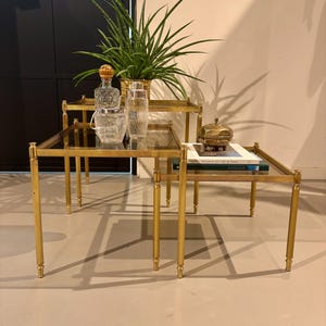 May include: A set of three nested tables featuring gold-toned metal frames and square glass tops. The tables are arranged in a tiered formation, with a green plant in a gold pot, a glass decanter, a glass ice bucket, and a book on the surfaces.