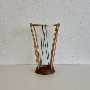 May include: A gold metal umbrella stand with red and black string accents. The stand has a round base and a curved top.