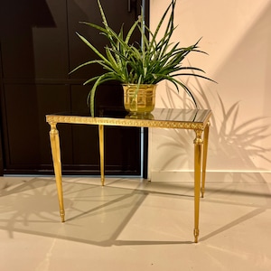 Peut inclure: Une table d'appoint carrée en or avec un plateau en verre noir et des détails ornés. La table a quatre pieds et repose sur un sol beige clair. Une plante verte en pot est posée sur la table.
