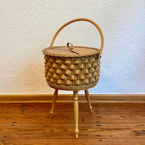 May include: A round, light brown woven basket with a textured, geometric pattern on the exterior. The basket features a flat lid with a small loop handle, a single arched carrying handle, and stands on three slender, light-colored wooden legs.