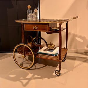 Può includere: Un carrello di servizio in legno con ringhiera in ottone, un cassetto con maniglia in ottone e due grandi ruote a raggi. Il carrello presenta un ripiano inferiore con una teiera in metallo, una piccola ciotola con coperchio e due libri.