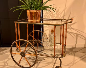 May include: A two-tiered serving trolley featuring a brass frame with bamboo-style accents, two large spoked wheels, and two smaller casters. The trolley has clear glass shelves, a handle, and holds a potted plant in a wooden basket and glassware.