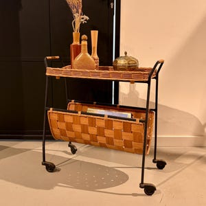 May include: A two-tiered trolley with a black metal frame and four castors. The top tier features a woven wicker tray, while the bottom tier consists of a deep, woven wooden basket. Three decorative vases and a lidded bowl sit on the top surface.