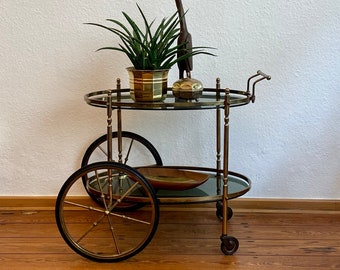 Puede incluir: Un carrito de bar de latón antiguo con dos estantes ovalados de vidrio y ruedas de radios. El carrito contiene una planta en maceta, una escultura de madera y un recipiente decorativo. El carrito está sobre un suelo de madera.