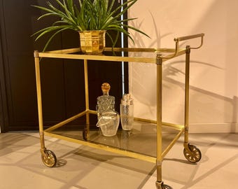 Op de afbeelding: Een goudkleurige barwagen met twee glazen planken en decoratieve wielen. De wagen bevat een potplant, een karaf, een cocktailshaker en een kleine ijsemmer. De wagen is ongeveer 76 cm hoog.