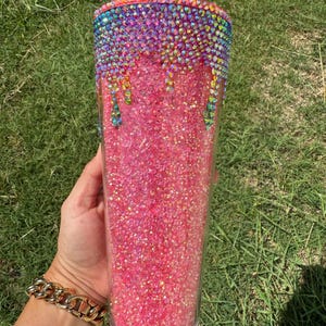 Puede incluir: Un vaso transparente lleno de purpurina rosa. La parte superior est&aacute; adornada con pedrer&iacute;a colorida que gotea por el lateral. Una pajita roja est&aacute; insertada en la parte superior. El vaso se sostiene sobre un fondo de hierba verde.