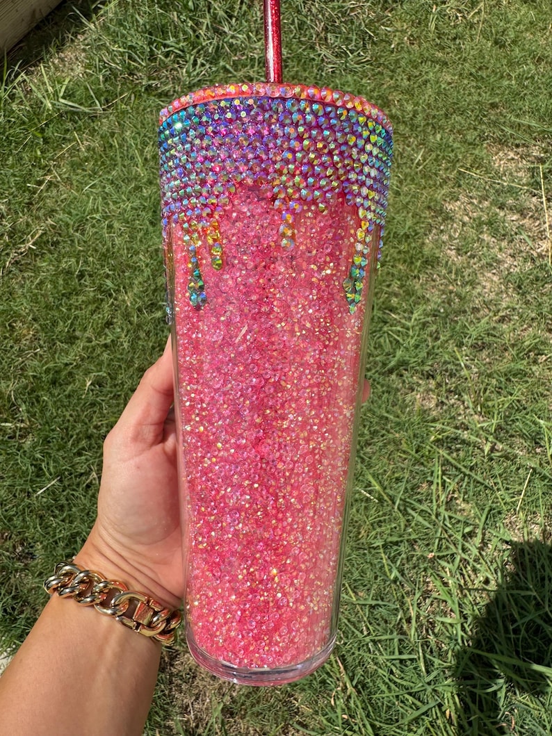 Puede incluir: Un vaso transparente lleno de purpurina rosa. La parte superior est&aacute; adornada con pedrer&iacute;a colorida que gotea por el lateral. Una pajita roja est&aacute; insertada en la parte superior. El vaso se sostiene sobre un fondo de hierba verde.