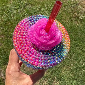 Puede incluir: Un vaso adornado con pedrer&iacute;a con una rosa rosa brillante y una pajita roja. La taza est&aacute; cubierta de pedrer&iacute;a iridiscente en un patr&oacute;n de arco&iacute;ris. La rosa es de color rosa brillante.