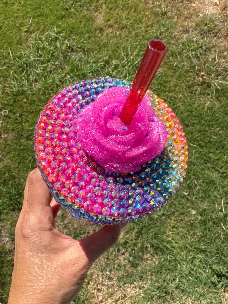 Puede incluir: Un vaso adornado con pedrer&iacute;a con una rosa rosa brillante y una pajita roja. La taza est&aacute; cubierta de pedrer&iacute;a iridiscente en un patr&oacute;n de arco&iacute;ris. La rosa es de color rosa brillante.
