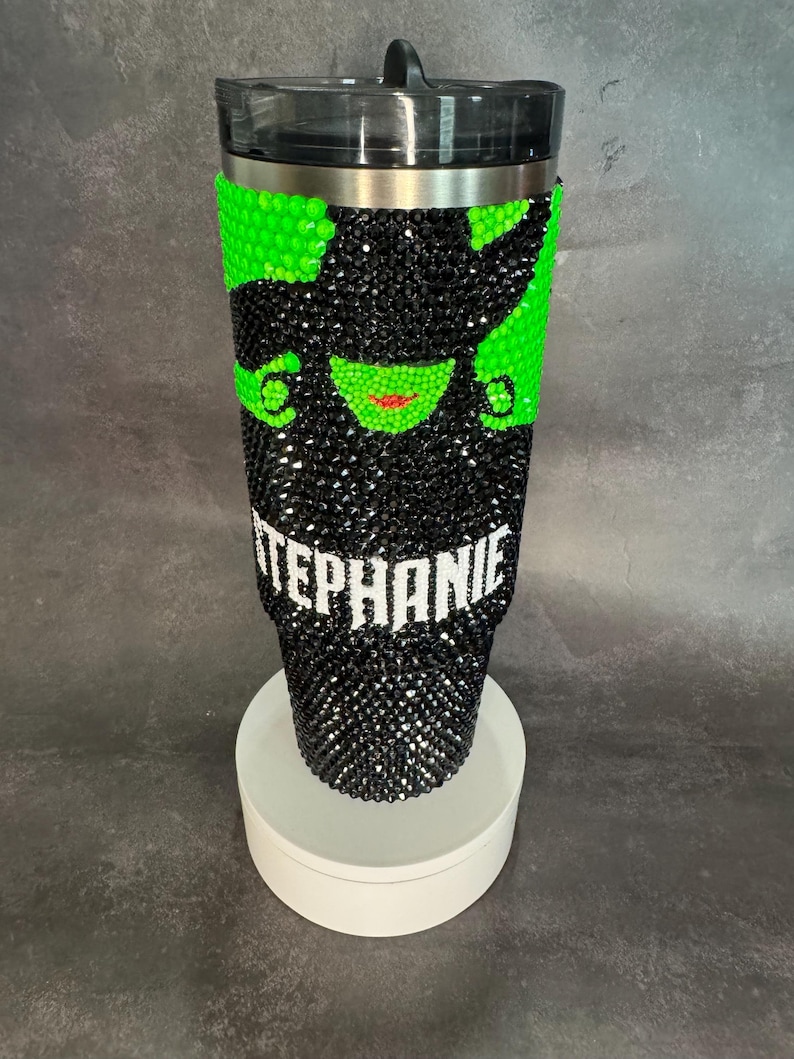May include: A black and green stainless steel tumbler with a rhinestone design of the Wicked Witch of the West. The tumbler has a black lid and a white name "STEPHANIE" on the bottom.
