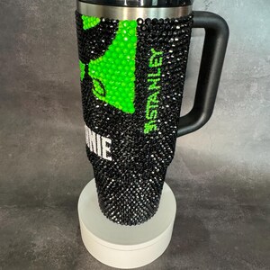 May include: A black and green stainless steel travel mug with a black handle and a clear lid. The mug is covered in rhinestones and has the word "Stanley" printed on the side. The mug is sitting on a white pedestal.