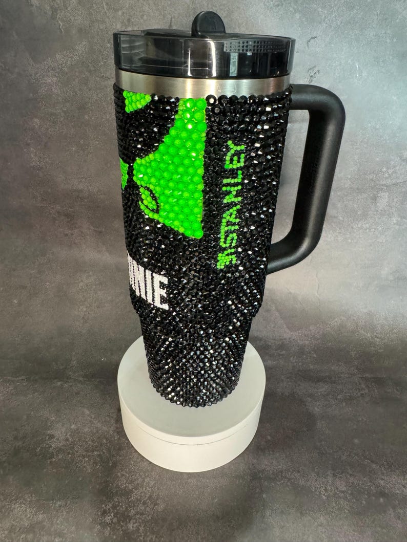 May include: A black and green stainless steel travel mug with a black handle and a clear lid. The mug is covered in rhinestones and has the word "Stanley" printed on the side. The mug is sitting on a white pedestal.