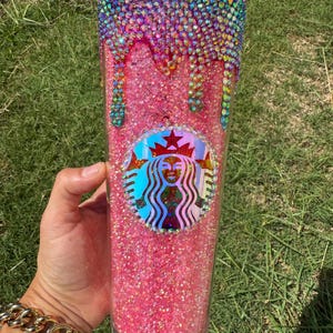 Puede incluir: Un vaso rosa brillante con el logotipo de Starbucks. La parte superior del vaso est&aacute; incrustada con pedrer&iacute;a colorida que gotea por el costado. El vaso lo sostiene una persona que lleva una pulsera dorada.