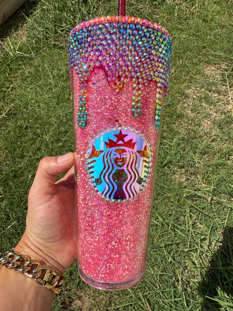 Puede incluir: Un vaso rosa brillante con el logotipo de Starbucks. La parte superior del vaso est&aacute; incrustada con pedrer&iacute;a colorida que gotea por el costado. El vaso lo sostiene una persona que lleva una pulsera dorada.