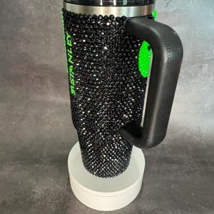 May include: Black travel mug with a black rhinestone design and a green inscription. The mug has a black handle and a black lid.