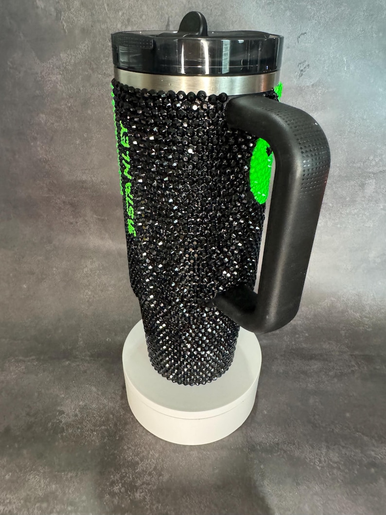 May include: Black travel mug with a black rhinestone design and a green inscription. The mug has a black handle and a black lid.