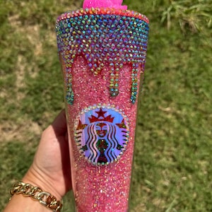 Puede incluir: Un vaso rosa brillante con pedrer&iacute;a iridiscente y una flor rosa en la parte superior. El borde superior est&aacute; adornado con una banda de pedrer&iacute;a colorida. El logotipo de Starbucks es visible en la parte delantera del vaso.