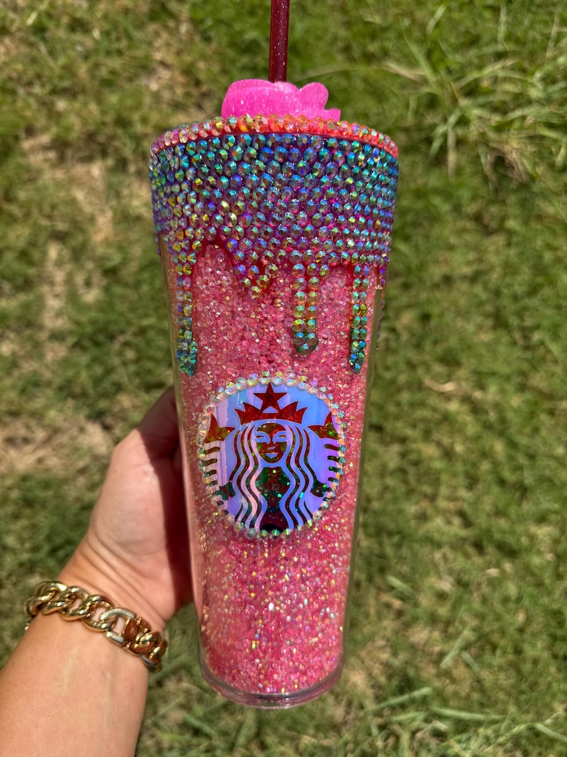 Puede incluir: Un vaso rosa brillante con pedrer&iacute;a iridiscente y una flor rosa en la parte superior. El borde superior est&aacute; adornado con una banda de pedrer&iacute;a colorida. El logotipo de Starbucks es visible en la parte delantera del vaso.