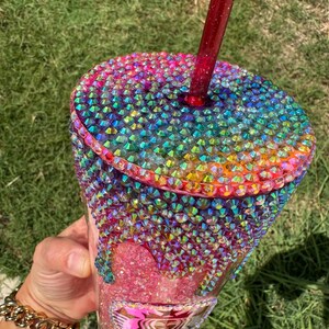 Puede incluir: Un vaso adornado con pedrer&iacute;a iridiscente en un arco&iacute;ris de colores. La tapa y la parte superior de la taza est&aacute;n cubiertas de pedrer&iacute;a. La pedrer&iacute;a gotea por el lateral del vaso. Una pajita roja est&aacute; insertada en la tapa.