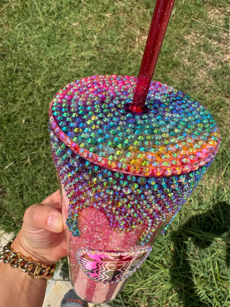 Puede incluir: Un vaso adornado con pedrer&iacute;a iridiscente en un arco&iacute;ris de colores. La tapa y la parte superior de la taza est&aacute;n cubiertas de pedrer&iacute;a. La pedrer&iacute;a gotea por el lateral del vaso. Una pajita roja est&aacute; insertada en la tapa.