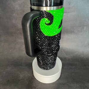 May include: A black and green travel mug with a handle and a lid. The mug is covered in rhinestones and has a green swirl design.