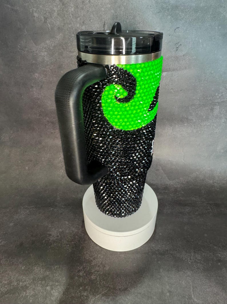 May include: A black and green travel mug with a handle and a lid. The mug is covered in rhinestones and has a green swirl design.