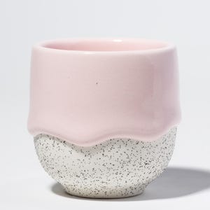 Könnte beinhalten: Eine kleine, handgefertigte Keramiktasse mit zweifarbigem Design. Die obere Hälfte ist einfarbig, glatt rosa, während die untere Hälfte eine gesprenkelte, strukturierte grau-weiße Oberfläche hat. Die Tasse hat eine abgerundete Form.