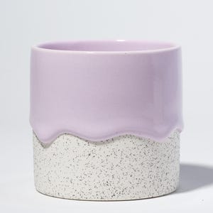 Op de afbeelding: Een keramische plantenbak met een tweekleurig ontwerp. De bovenste helft is effen lavendelkleurig en de onderste helft is bespikkeld met grijze stippen. De twee secties worden gescheiden door een golvende lijn.