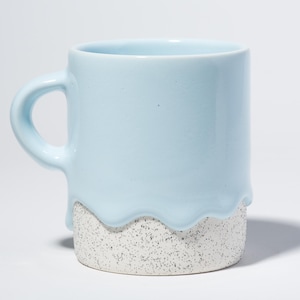 May include: A light blue ceramic mug with a distinctive design. The top half of the mug is a solid light blue, while the bottom half has a speckled, textured appearance. The light blue paint appears to be dripping down the side of the mug.