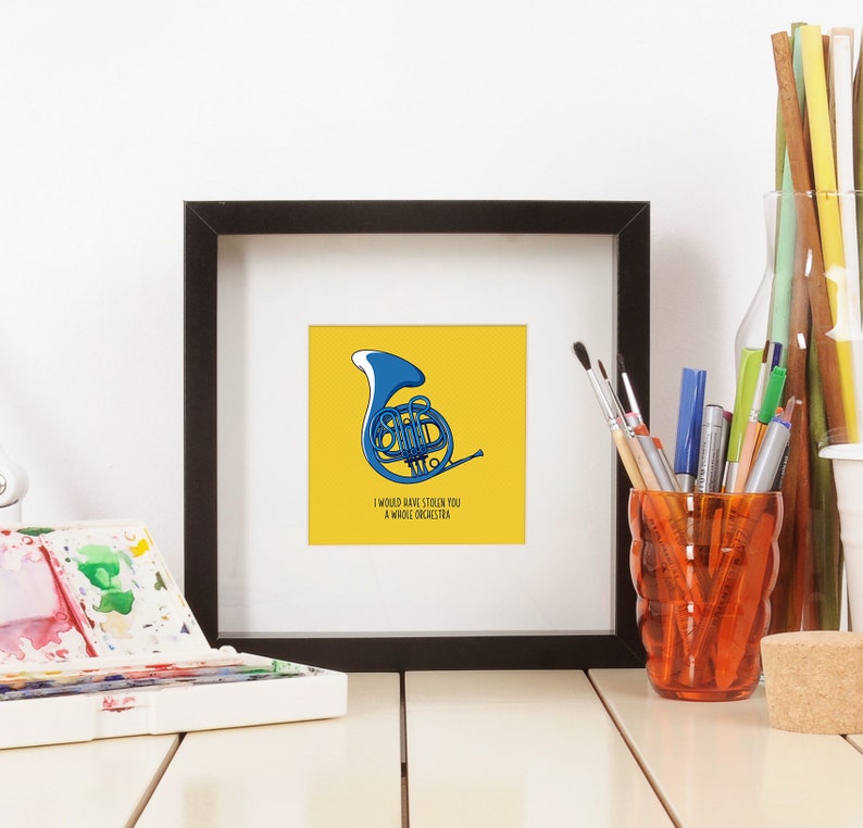 Op de afbeelding: Een ingelijste kunstprint met een blauwe Franse hoorn illustratie op een gele achtergrond. De tekst "I would have stolen you a whole orchestra" staat eronder. De zwarte lijst contrasteert met het kunstwerk, dat op een wit oppervlak wordt getoond.