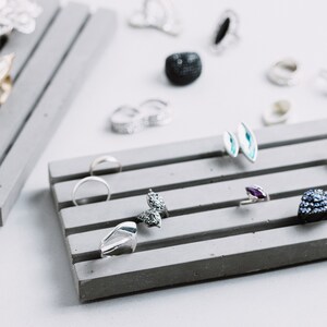 May include: A gray concrete ring holder with five slots for rings and earrings. The holder is filled with various silver, blue, and purple rings and earrings.