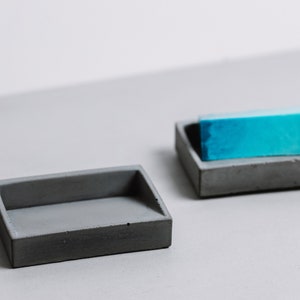 May include: Two gray concrete soap dishes. One dish is empty and the other holds a light blue bar of soap.