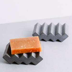 May include: Two gray concrete soap dishes with a zig-zag pattern. One dish has a bar of orange soap on top.