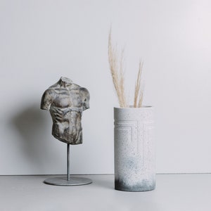 Puede incluir: Un jarrón de cerámica blanco con una superficie texturizada y un diseño geométrico. El jarrón está lleno de hierbas secas beige y está sentado sobre una superficie gris junto a una escultura de yeso de un torso.