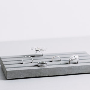 May include: A gray concrete ring holder with five slots for rings. The holder has a textured surface and is empty except for four silver rings.