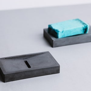 May include: Two gray concrete soap dishes. One dish is empty and the other has a light blue bar of soap on top.