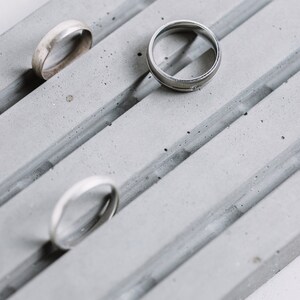 May include: Three silver rings on a gray concrete ring holder. The rings are different sizes and styles.