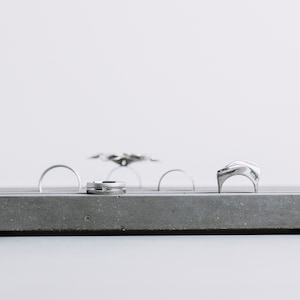 May include: A gray concrete ring holder with five slots for rings. The holder is on a white surface. There are four silver rings and one silver ring with a unique design.