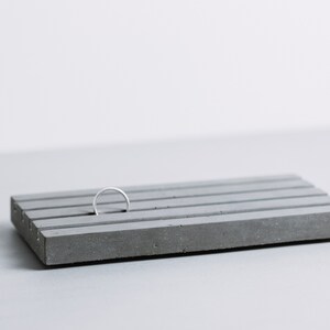 May include: A gray concrete ring holder with five grooves. A silver ring is placed in one of the grooves.