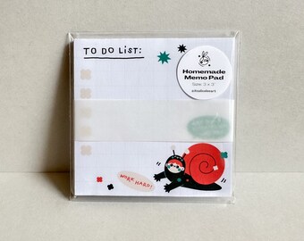May include: A white memo pad with a snail illustration and the text "TO DO LIST: WORK HARD!"  The memo pad is packaged in a clear plastic sleeve and has a sticker that says "Homemade Memo Pad Size: 3 x 3 aitsaliceleeart"