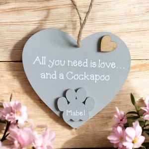 May include: A gray wooden heart-shaped sign with the text "All you need is love... and a Cockapoo" and a gray paw print with the name "Mabel" written on it. A small wooden heart is attached to the top of the sign.