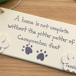 May include: White wooden sign with the text "A home is not complete without the pitter patter of Cavapoochon feet". Two white paw prints with the names "Harley" and "Jessie" are attached to the sign.