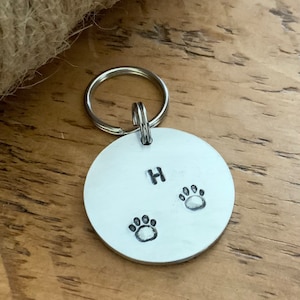May include: Silver dog tag with a paw print design and the letter H engraved on it.