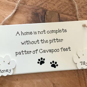 May include: White wooden sign with the text "A home is not complete without the pitter patter of Cavapoo feet". Two white paw prints are attached to the sign, one says "Honey" and the other says "Tilly".