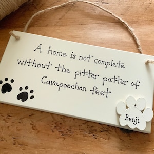 May include: White wooden sign with black text that reads "A home is not complete without the pitter patter of Cavapoocho feet".  The sign has two black paw prints and a white paw print with the name "Benji" written on it.