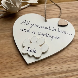 May include: Cream-colored heart-shaped wooden plaque with the text "All you need is love... and a Cockapoo." Features a paw print with the name "Rolo" and a small wooden heart. A jute string is attached for hanging.