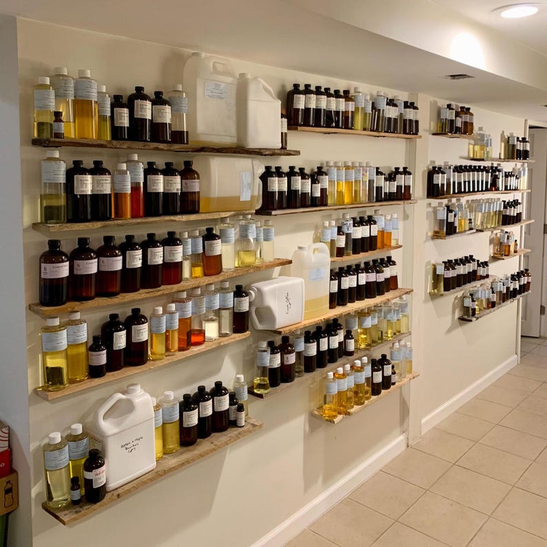 May include: A wall-mounted display featuring multiple rows of wooden shelves holding numerous glass and plastic bottles filled with various colored liquids, including amber, yellow, and clear oils, along with several large white plastic jugs.