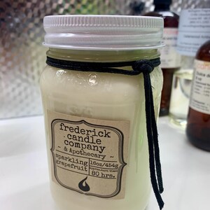 May include: A clear glass mason jar candle filled with off-white wax, topped with a white lid. A black cord is tied around the neck. The label reads: frederick candle company & Apothecary, sparkling grapefruit, 16oz/454g, approx. burn time: 80 hrs.