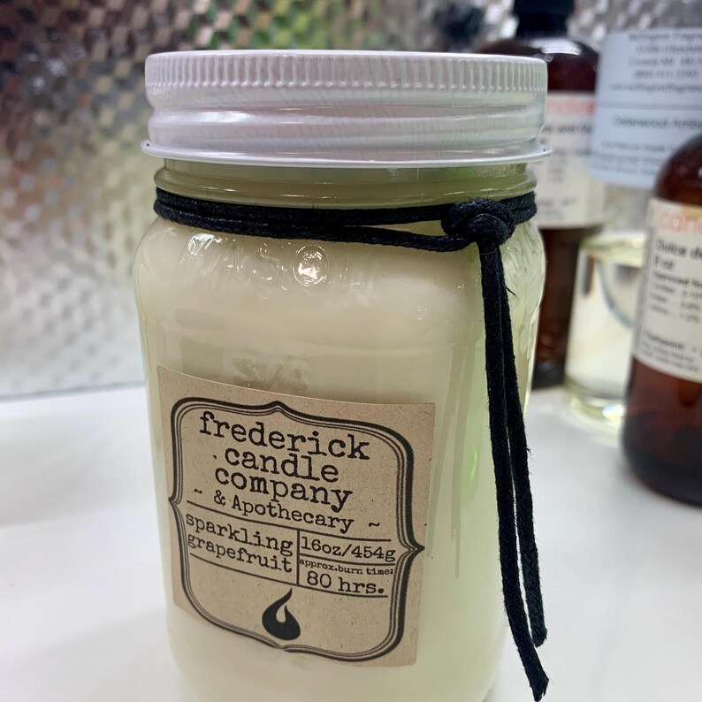 May include: A clear glass mason jar candle filled with off-white wax, topped with a white lid. A black cord is tied around the neck. The label reads: frederick candle company & Apothecary, sparkling grapefruit, 16oz/454g, approx. burn time: 80 hrs.